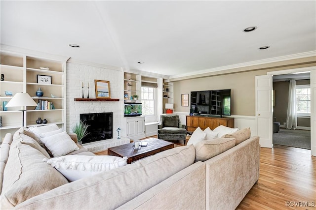 living area featuring crown molding, a brick fireplace, light wood-type flooring, built in shelves, and a wainscoted wall