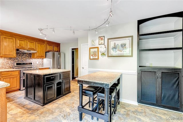 kitchen with appliances with stainless steel finishes, light stone counters, backsplash, under cabinet range hood, and a kitchen island