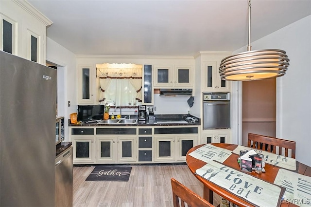 kitchen with appliances with stainless steel finishes, dark countertops, light wood-type flooring, a sink, and under cabinet range hood