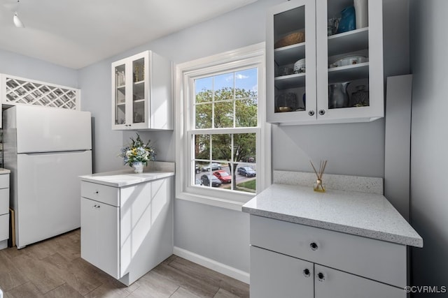 kitchen featuring freestanding refrigerator, white cabinets, light stone counters, glass insert cabinets, and baseboards