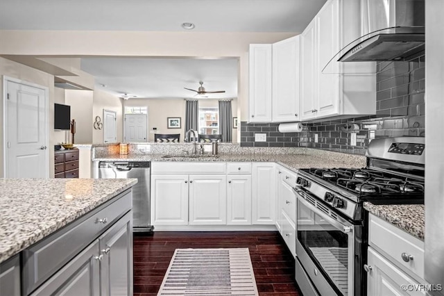 kitchen featuring wall chimney range hood, appliances with stainless steel finishes, a sink, a peninsula, and dark wood-style floors