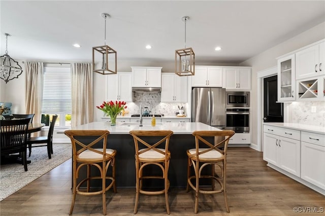 kitchen with appliances with stainless steel finishes, light countertops, tasteful backsplash, under cabinet range hood, and a center island with sink