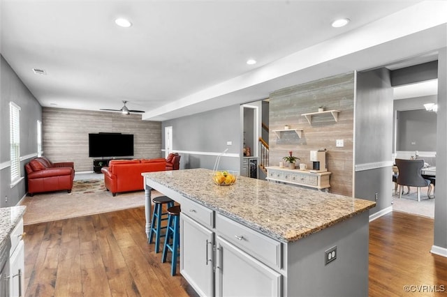 kitchen with ceiling fan, light stone countertops, a kitchen breakfast bar, an accent wall, and dark wood-style floors