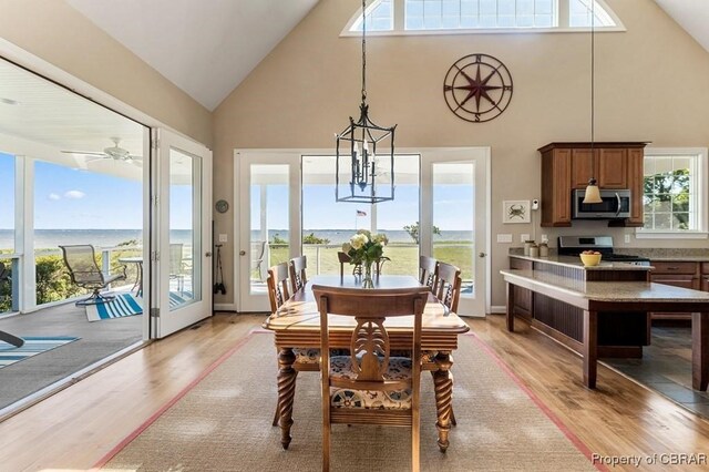 dining space featuring ceiling fan with notable chandelier, high vaulted ceiling, a water view, and light wood finished floors