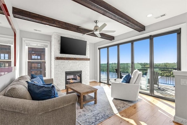 living room featuring a ceiling fan, a stone fireplace, visible vents, beamed ceiling, and light wood-style floors