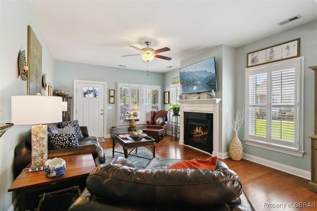 living room with visible vents, wood finished floors, baseboards, a warm lit fireplace, and a ceiling fan