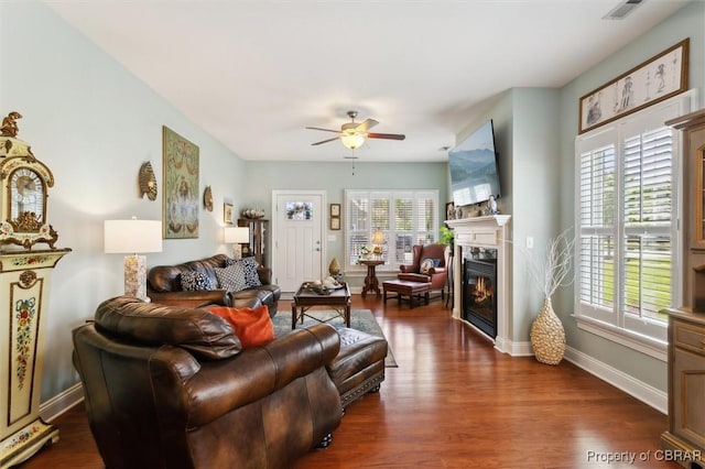 living area featuring dark wood-style flooring, a glass covered fireplace, a ceiling fan, baseboards, and visible vents