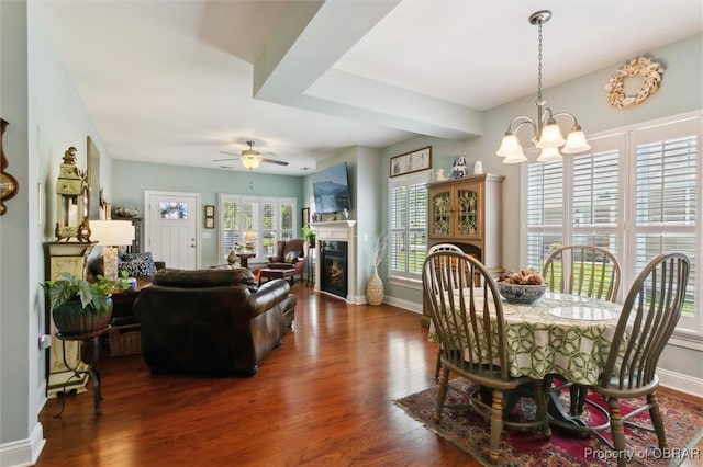 dining space with baseboards, a glass covered fireplace, dark wood-style floors, and ceiling fan with notable chandelier