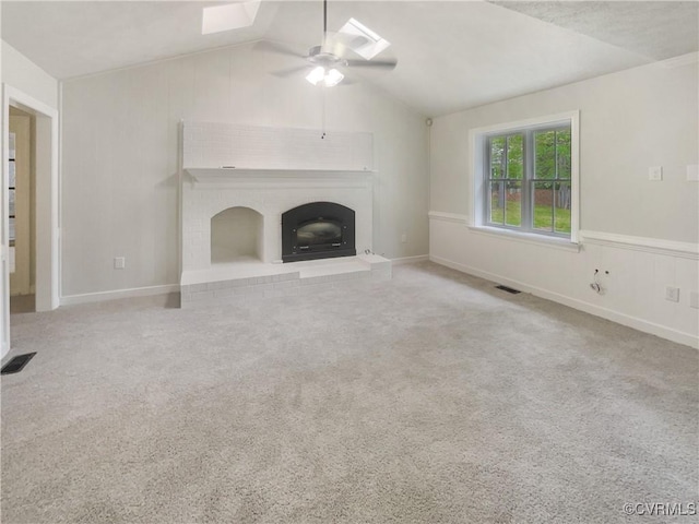 unfurnished living room featuring ceiling fan, vaulted ceiling with skylight, a brick fireplace, carpet, and visible vents