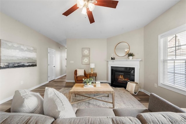 living area featuring ceiling fan, a glass covered fireplace, wood finished floors, and baseboards
