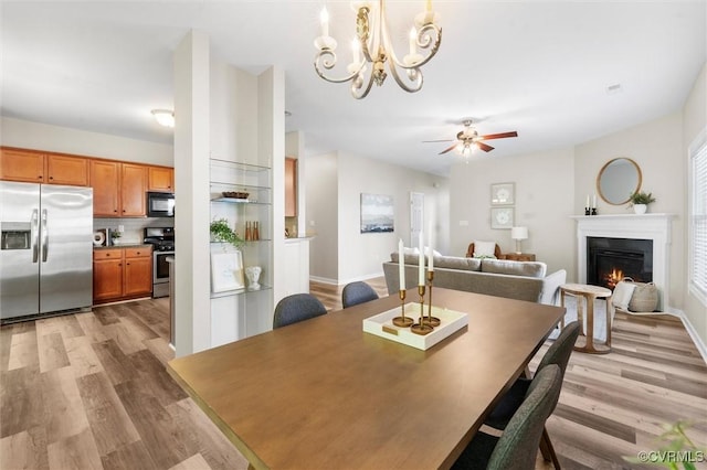 dining area featuring light wood-style flooring, a lit fireplace, a ceiling fan, a chandelier, and baseboards