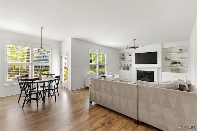 living area featuring built in features, a glass covered fireplace, wood finished floors, baseboards, and a chandelier