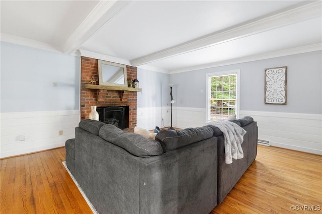 living room with beamed ceiling, light wood finished floors, wainscoting, a brick fireplace, and crown molding