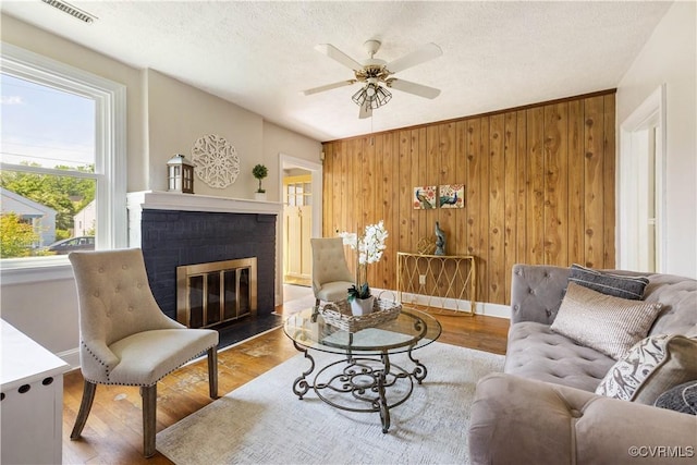living area featuring wood finished floors, a ceiling fan, a brick fireplace, wooden walls, and a textured ceiling