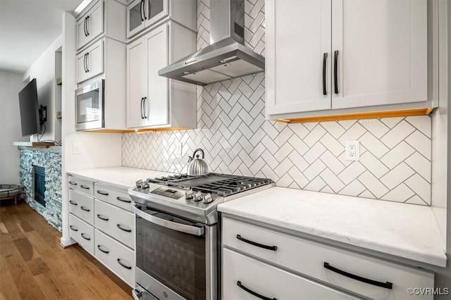 kitchen with wall chimney range hood, appliances with stainless steel finishes, light stone countertops, light wood finished floors, and glass insert cabinets