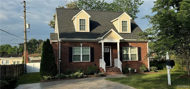 cape cod house featuring roof with shingles, fence, a front lawn, and brick siding