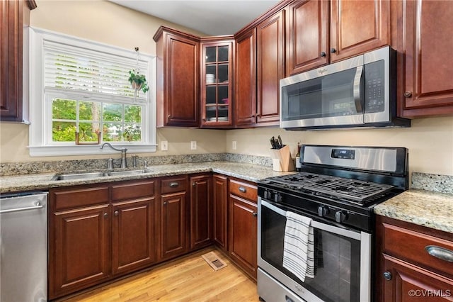 kitchen featuring appliances with stainless steel finishes, a sink, light wood finished floors, glass insert cabinets, and light stone counters