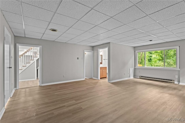 unfurnished living room featuring a baseboard heating unit, baseboards, light wood-style floors, a paneled ceiling, and stairway