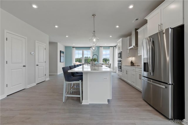 kitchen with appliances with stainless steel finishes, a sink, wall chimney range hood, a kitchen island with sink, and white cabinetry