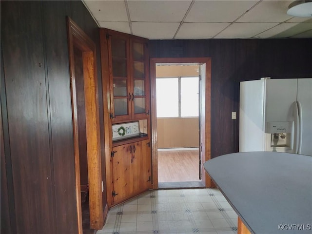 kitchen featuring white refrigerator with ice dispenser, light flooring, a drop ceiling, glass insert cabinets, and wooden walls
