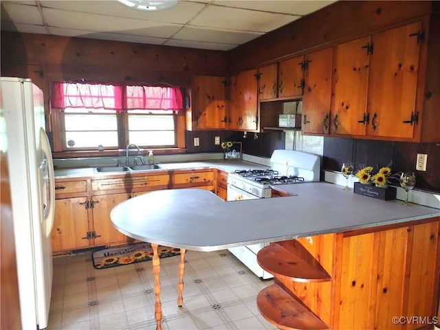kitchen with white appliances, a sink, light flooring, a peninsula, and a drop ceiling