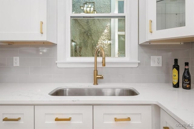kitchen view of a sink, white cabinets, decorative backsplash, glass insert cabinets, and light stone counters