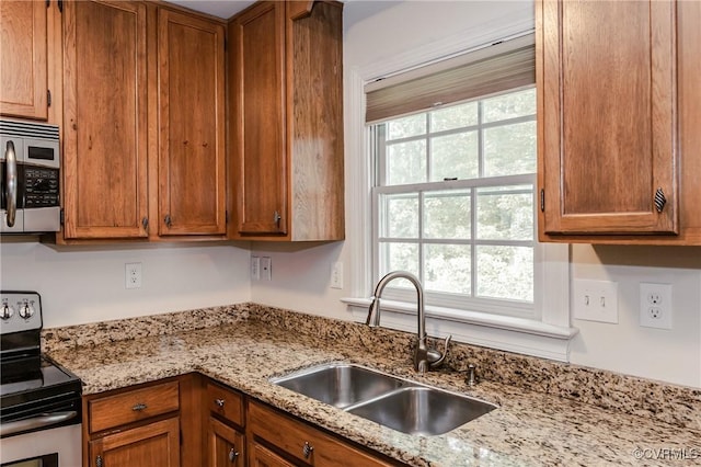 kitchen featuring stainless steel appliances, a sink, brown cabinetry, and light stone countertops