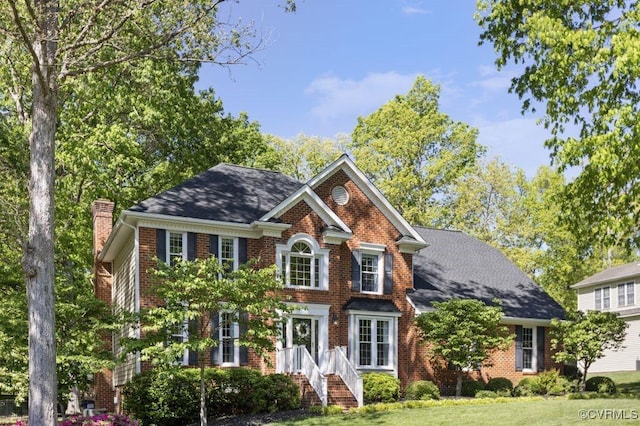 georgian-style home with a chimney, brick siding, a front yard, and a shingled roof