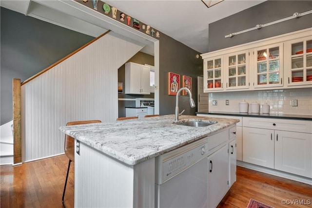kitchen with white dishwasher, a sink, wood finished floors, an island with sink, and glass insert cabinets