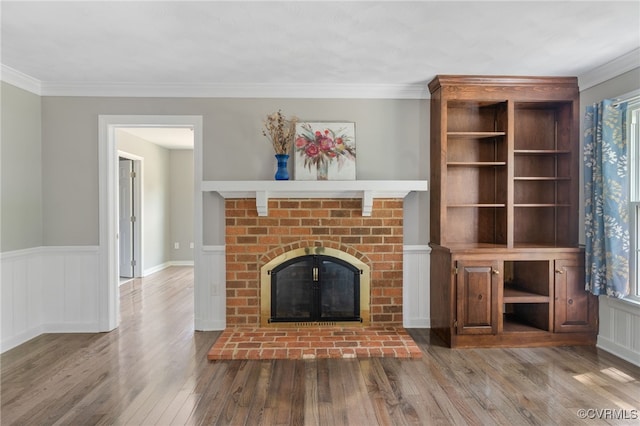 unfurnished living room featuring a brick fireplace, wood finished floors, ornamental molding, and wainscoting