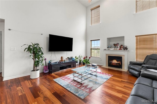 living area featuring a towering ceiling, a fireplace with flush hearth, wood finished floors, and baseboards