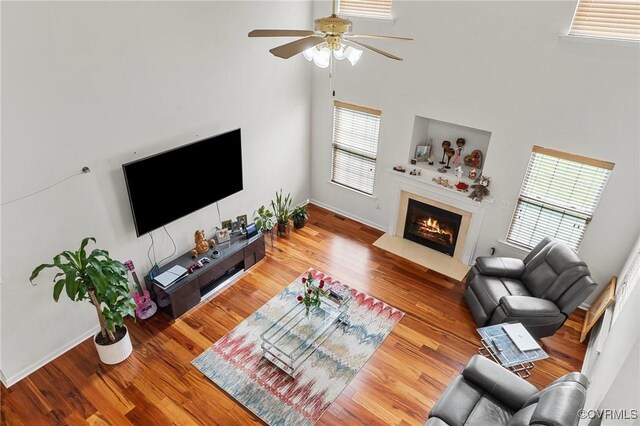 living area featuring a high ceiling, a fireplace with flush hearth, plenty of natural light, a ceiling fan, and wood finished floors