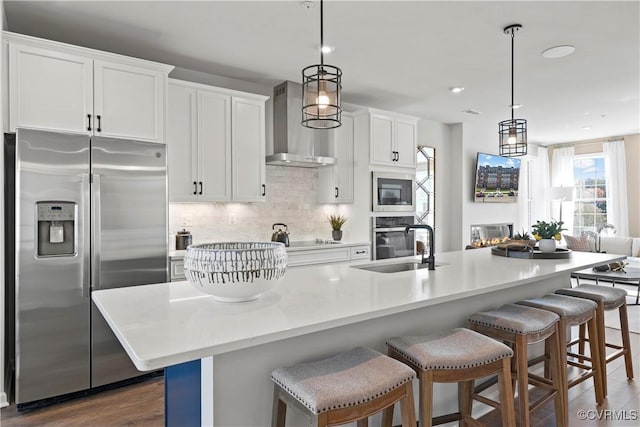 kitchen featuring appliances with stainless steel finishes, wall chimney range hood, an island with sink, backsplash, and white cabinets