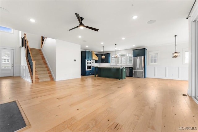 unfurnished living room with a decorative wall, a ceiling fan, wainscoting, light wood finished floors, and healthy amount of natural light