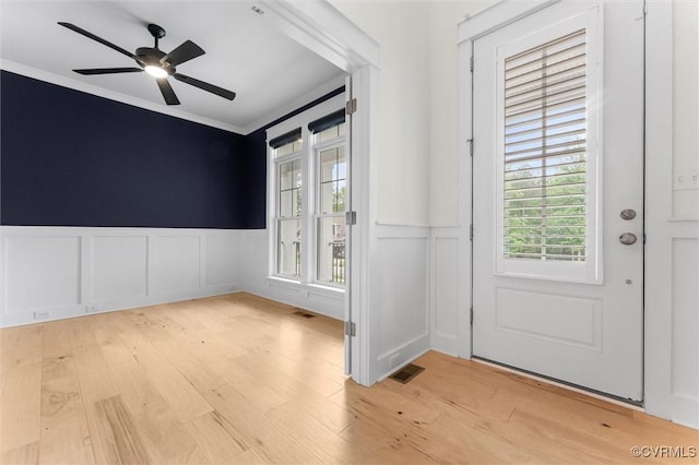 entrance foyer with ornamental molding, a decorative wall, wainscoting, light wood-style floors, and a ceiling fan