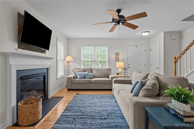 living area featuring stairway, light wood-type flooring, ceiling fan, a fireplace with flush hearth, and baseboards