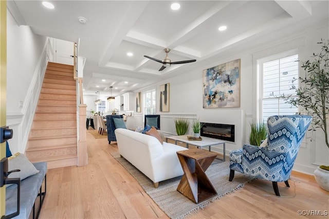 living area featuring stairway, light wood-type flooring, coffered ceiling, a glass covered fireplace, and beamed ceiling