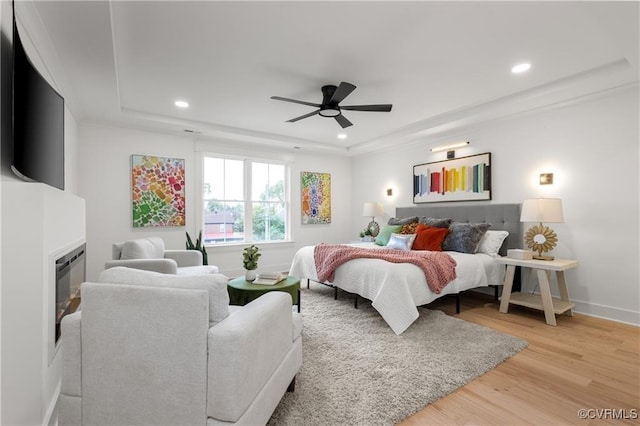 bedroom with light wood-type flooring, recessed lighting, a tray ceiling, baseboards, and a glass covered fireplace