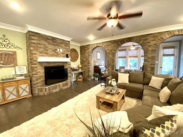 living room with arched walkways, wood finished floors, a ceiling fan, a brick fireplace, and ornamental molding