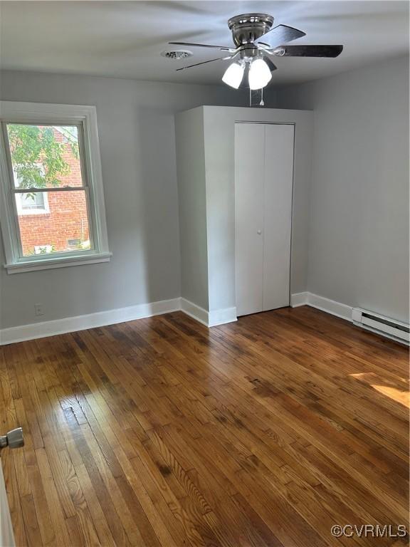 unfurnished bedroom with a baseboard heating unit, hardwood / wood-style floors, baseboards, a ceiling fan, and a closet