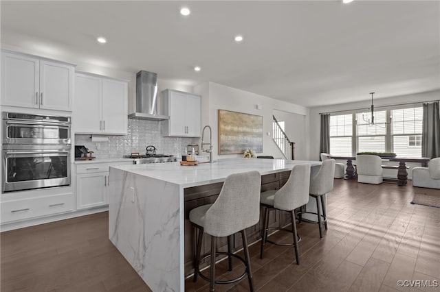 10109 Hollythorne Ln, Mechanicsville, VA 23116 - kitchen with wall chimney range hood, double oven, a kitchen island with sink, backsplash, and a kitchen bar
