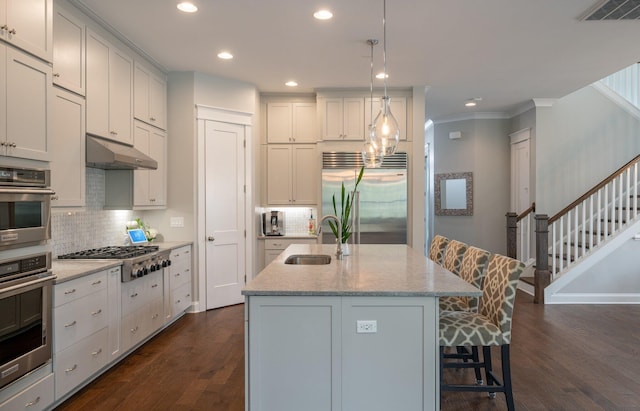 kitchen with sink, a kitchen island with sink, stainless steel appliances, light stone counters, and decorative light fixtures