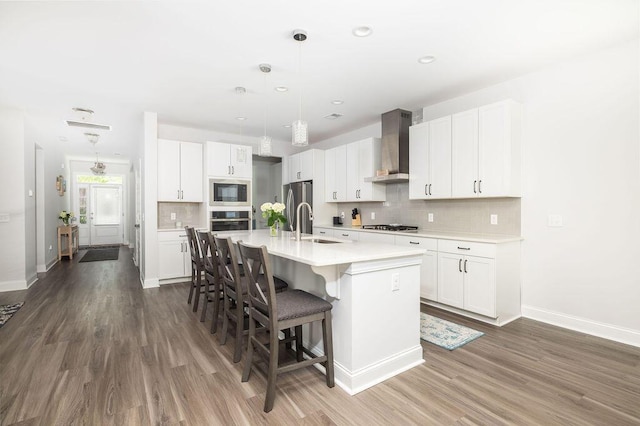 kitchen featuring an island with sink, light countertops, wall chimney exhaust hood, a kitchen bar, and appliances with stainless steel finishes