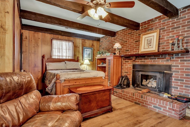 bedroom with ceiling fan, a brick fireplace, beamed ceiling, light hardwood / wood-style floors, and wooden walls