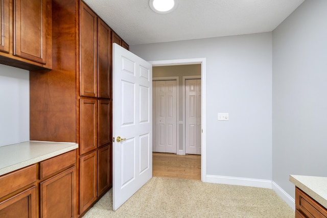 kitchen with brown cabinetry, a textured ceiling, light colored carpet, light countertops, and baseboards