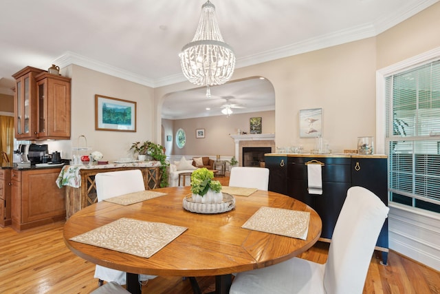 dining space featuring light wood finished floors, arched walkways, ornamental molding, a fireplace, and ceiling fan with notable chandelier