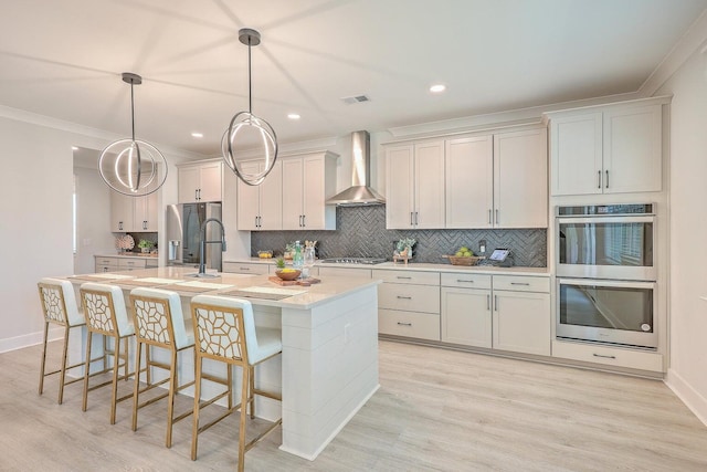 kitchen with wall chimney range hood, a center island with sink, decorative light fixtures, appliances with stainless steel finishes, and light hardwood / wood-style floors