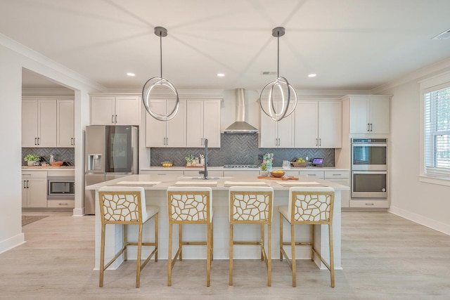 kitchen featuring wall chimney range hood, hanging light fixtures, a center island with sink, appliances with stainless steel finishes, and a breakfast bar