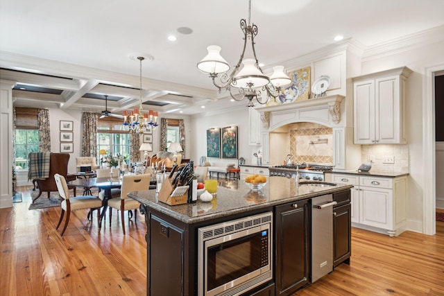 kitchen featuring coffered ceiling, stainless steel microwave, a kitchen island with sink, light wood-style floors, and a notable chandelier