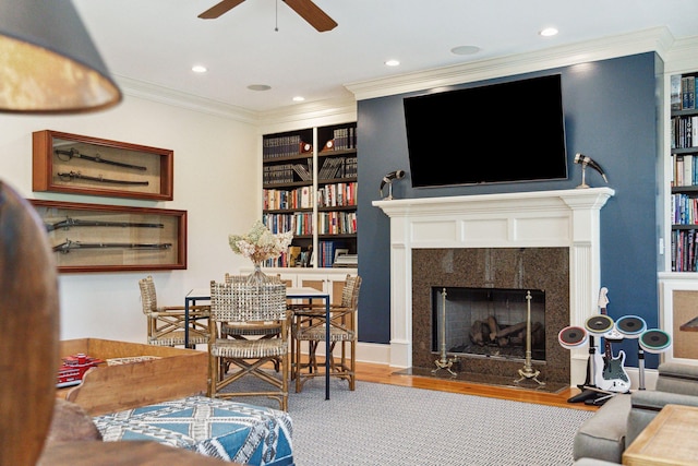 living room featuring built in features, a fireplace, crown molding, a ceiling fan, and wood finished floors